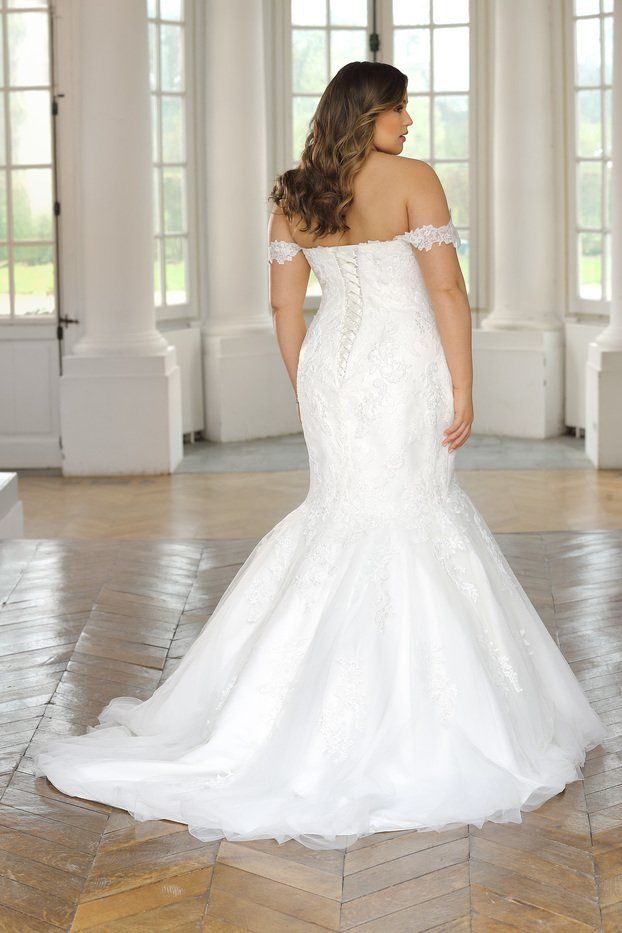 Una sposa vista di spalle, con indosso un abito da sposa bianco a sirena, con spalle scoperte e strascico, in una stanza luminosa.