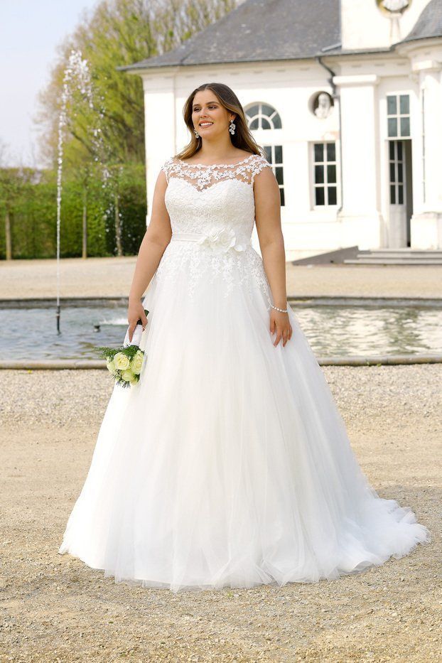 Una persona sorridente con indosso un abito da sposa di pizzo bianco, che tiene in mano un piccolo bouquet, è all'aperto davanti a un edificio e a una fontana.