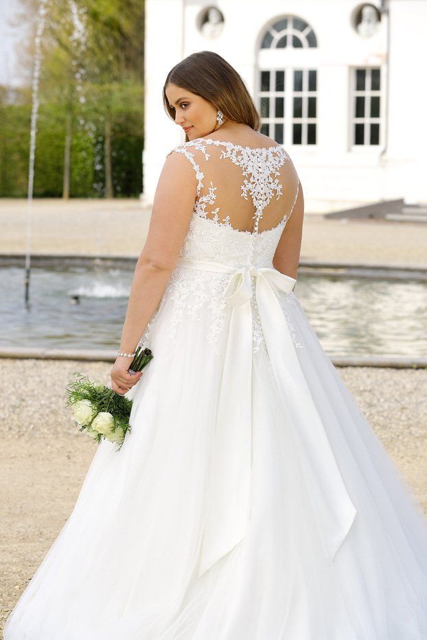 Una sposa con un abito bianco a trapezio con schiena in pizzo tiene in mano un bouquet di rose bianche davanti a un edificio con una fontana.