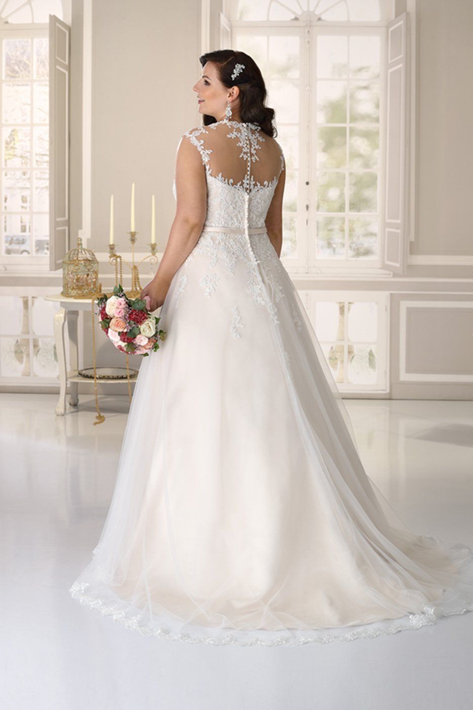 Una sposa in un abito da sposa bianco di pizzo lungo fino ai piedi, con schiena trasparente, tiene in mano un bouquet in una stanza luminosa ed elegante.