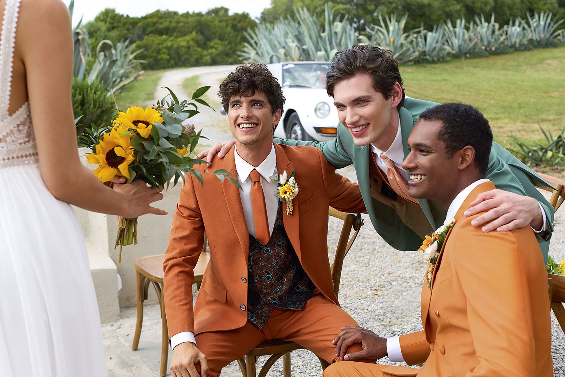 Una sposa che tiene in mano dei girasoli guarda verso tre persone che indossano abiti colorati, sedute all'aperto vicino a un'auto d'epoca.