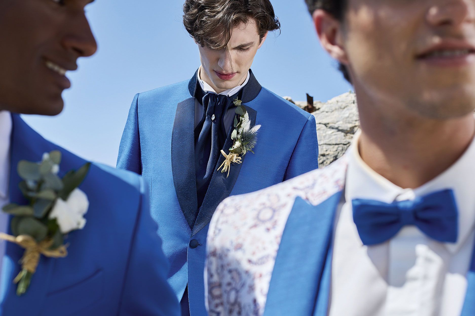 Tre persone che indossano abiti da sposo blu e boutonnière, con una persona al centro che guarda verso il basso.