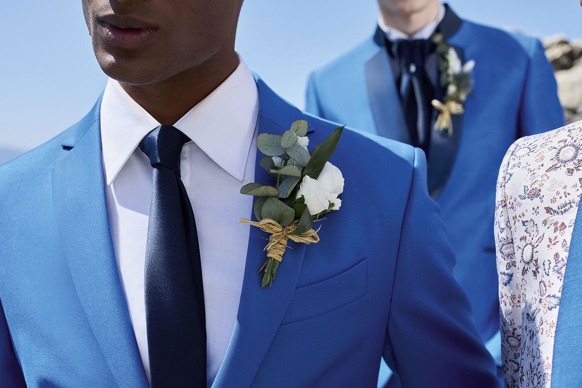 Tre individui che indossano abiti blu brillante con camicie bianche, cravatte scure e boutonnière floreali.