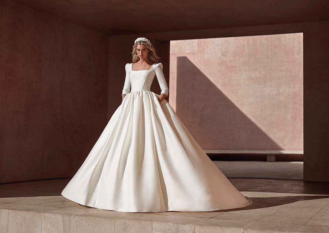 Una modella indossa un abito da sposa a maniche lunghe, con scollo quadrato e gonna ampia, in piedi sotto un arco di pietra minimalista.