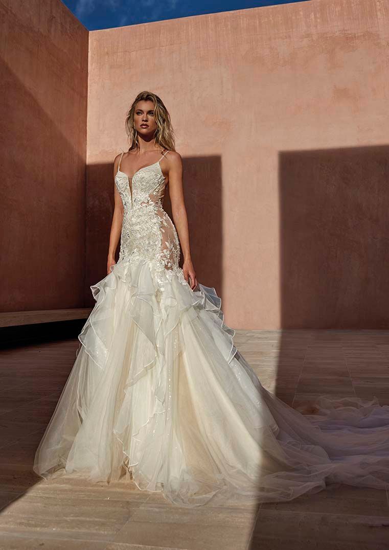 Una modella indossa un elaborato abito da sposa lungo fino ai piedi in pizzo bianco, con una gonna a strati di tulle, in un'ambientazione desertica all'aperto.