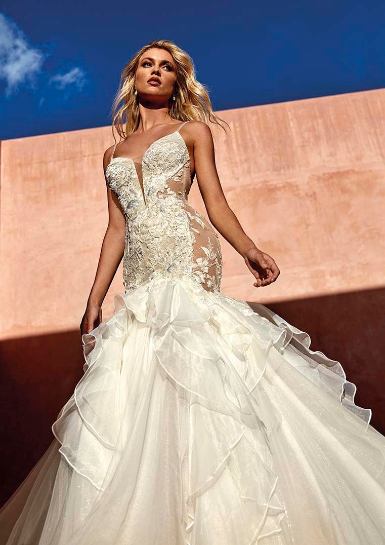 Una persona indossa un elaborato abito da sposa di pizzo bianco con una profonda scollatura a V e un'ampia gonna di tulle a strati e balze.