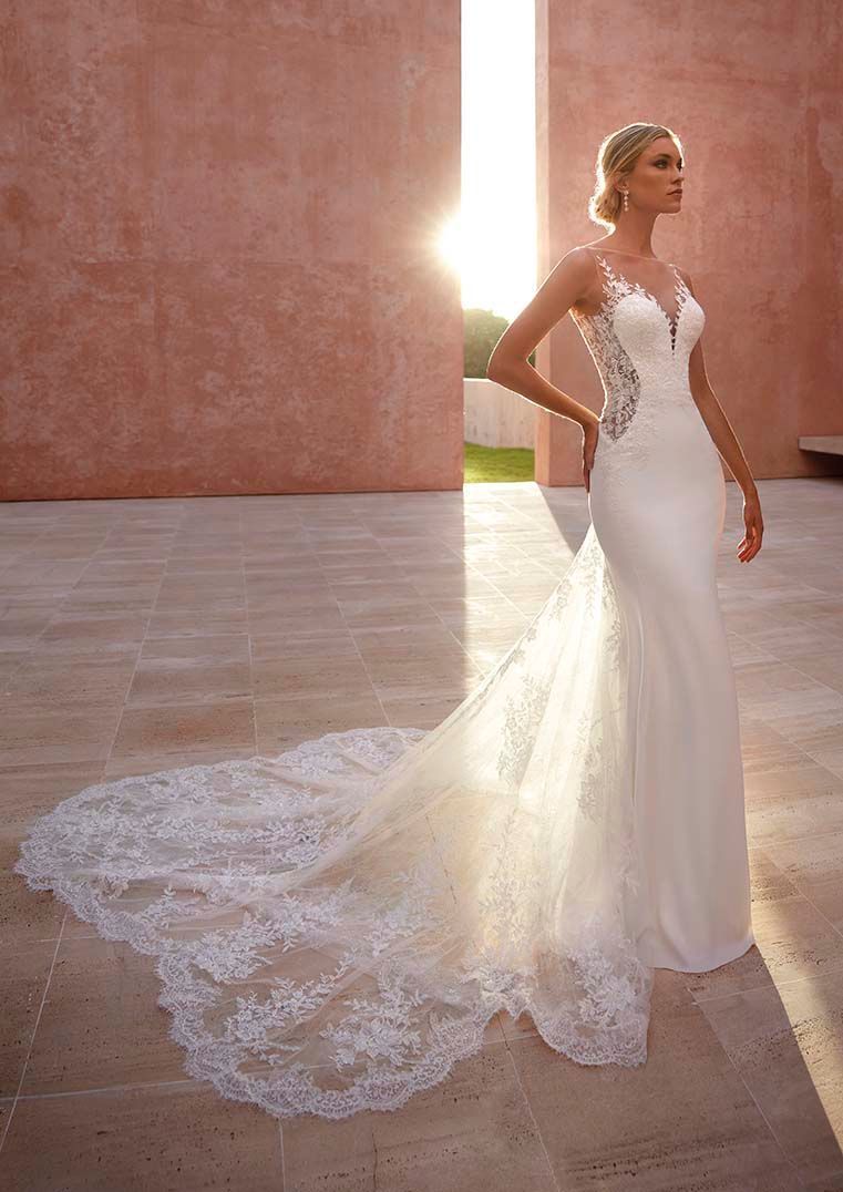 Una persona indossa un abito da sposa bianco aderente con corpetto e lungo strascico di pizzo, in piedi davanti a un muro al tramonto.
