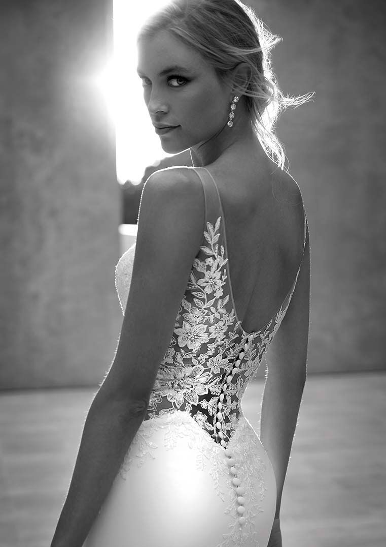 Una persona con indosso un abito da sposa senza schienale, impreziosito da dettagli in pizzo e con una fila di bottoni, si volta indietro all'aperto.