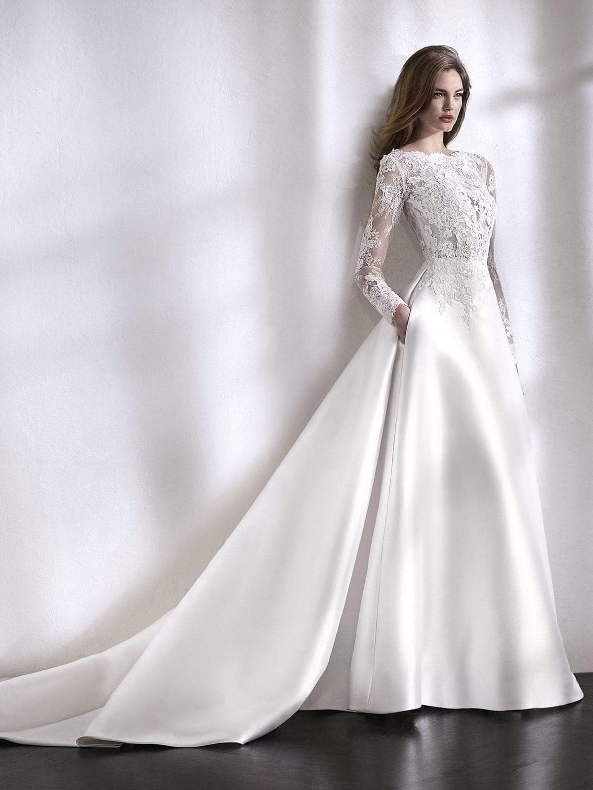 Abito da sposa a maniche lunghe con corpetto in pizzo e gonna lunga in raso liscio con strascico, indossato da una modella.