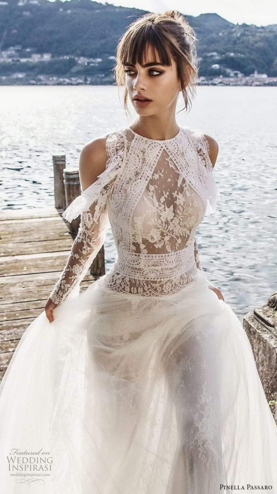 Una persona che indossa un elaborato abito da sposa di pizzo bianco con gonna di tulle è in piedi su un pontile di legno in riva a un lago tranquillo.