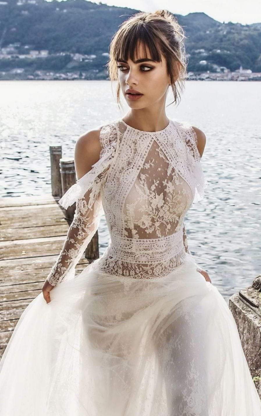 Una modella, con indosso un elaborato abito da sposa bianco a maniche lunghe in pizzo, posa su un pontile di legno vicino a un lago.