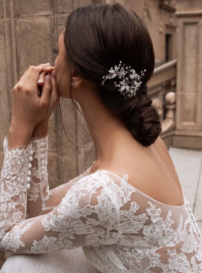 Una persona con un abito da sposa di pizzo, i capelli raccolti in uno chignon basso e un fermaglio decorativo, vista di profilo sullo sfondo di un edificio in pietra.