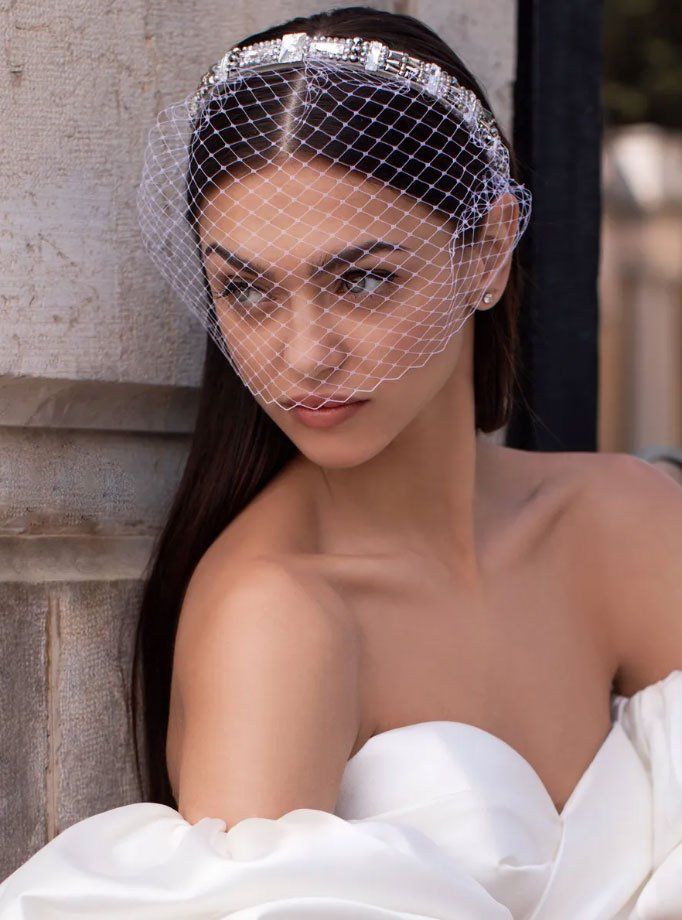 Una persona con lunghi capelli scuri che indossa una fascia per capelli decorata con cristalli e un velo a gabbia sopra un abito bianco a spalle scoperte.