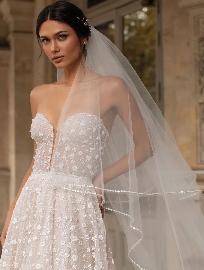 Una modella indossa un abito da sposa a trapezio senza spalline, impreziosito da decorazioni floreali, con una profonda scollatura e un velo trasparente.