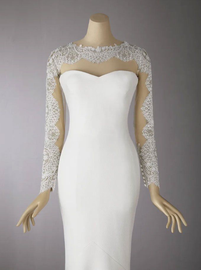 Un abito da sposa bianco lungo fino ai piedi, con scollo a cuore e maniche lunghe in pizzo trasparente, indossato da un manichino.