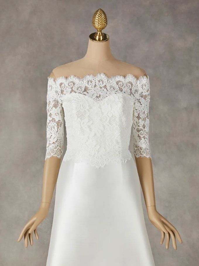 Un abito da sposa bianco, con scollo a spalle scoperte, corpetto in pizzo e maniche a tre quarti in pizzo, esposto su un manichino.