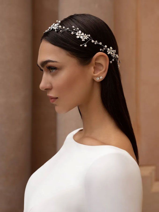Ritratto di profilo di una persona con i capelli scuri che indossa un delicato copricapo floreale e di cristalli, abbinata a un semplice abito bianco.