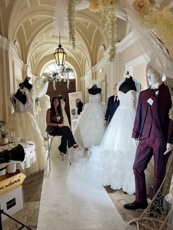 Una persona siede in un corridoio pieno di abiti da sposa su manichini e un abito formale, decorato con tessuto bianco.