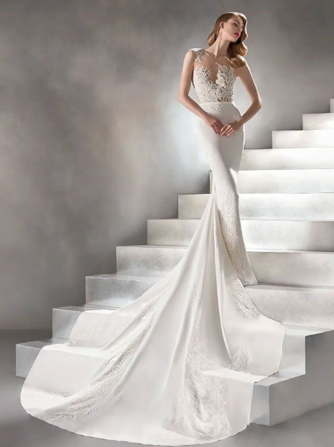 Una modella indossa un abito da sposa bianco aderente, con dettagli in pizzo e un lungo strascico, in posa su una scalinata.
