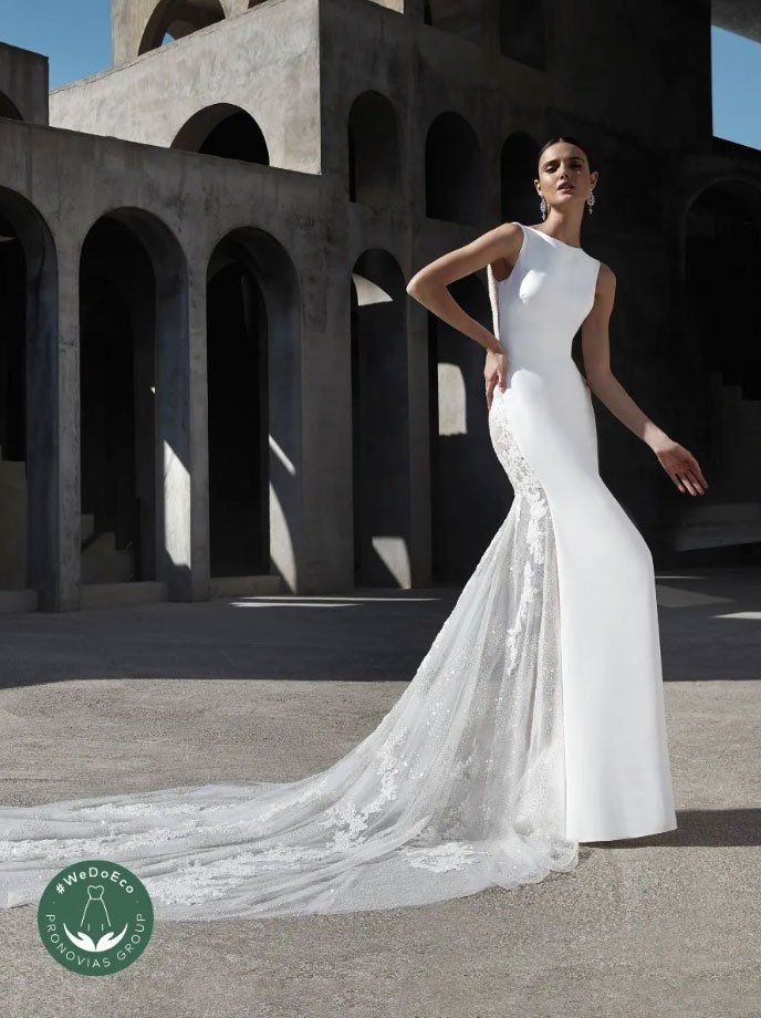Una modella indossa un abito da sposa a sirena bianco e aderente, con inserti laterali in pizzo e un lungo strascico, sullo sfondo di archi.