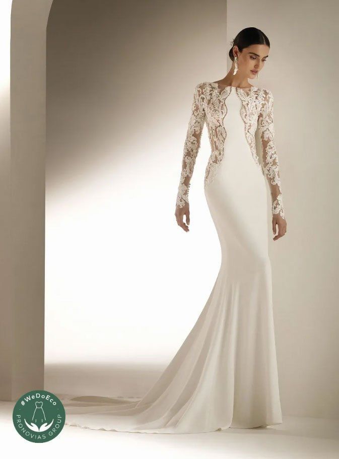 Una modella indossa un abito da sposa bianco a maniche lunghe, lungo fino ai piedi, con corpetto in pizzo, su uno sfondo minimalista.
