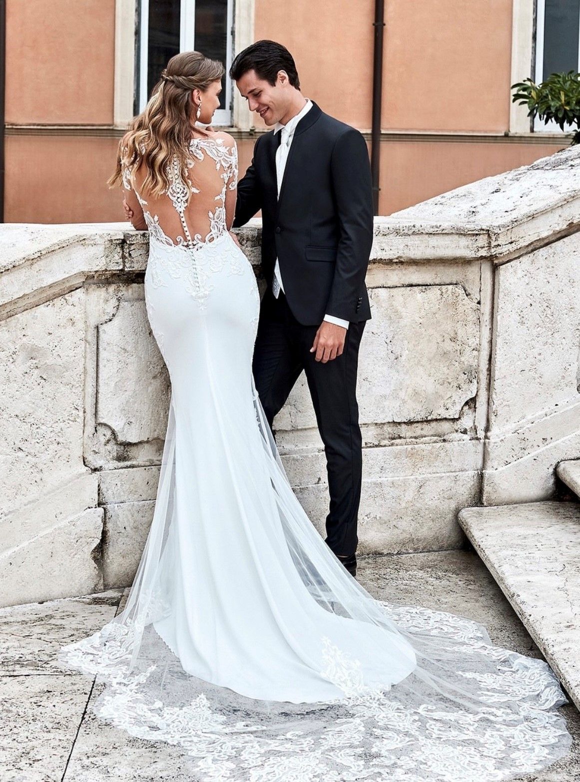 Una coppia posa su una scalinata di pietra; una persona con un abito da sposa bianco con schiena scoperta in pizzo è girata di spalle e guarda una persona in abito elegante.