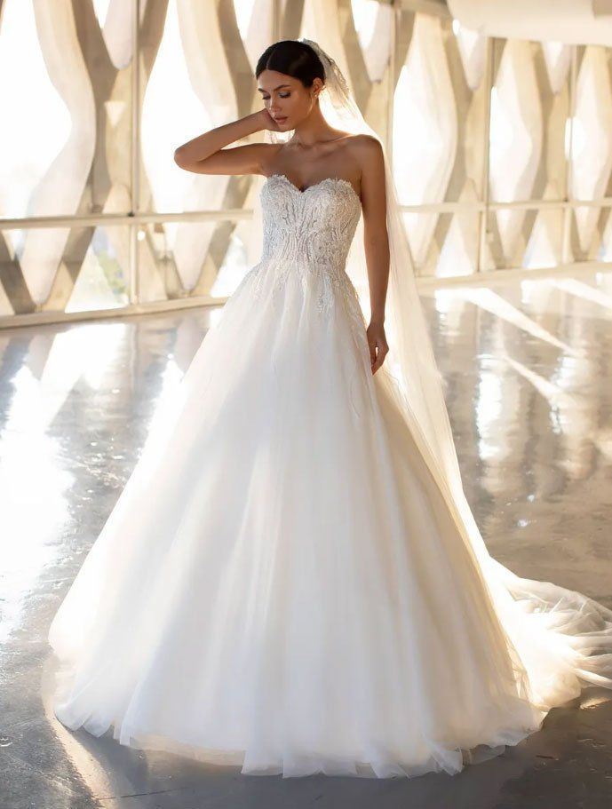 Una sposa in un abito da ballo senza spalline con corpetto di pizzo, ampia gonna di tulle e lungo velo, è in piedi in un corridoio luminoso e moderno.