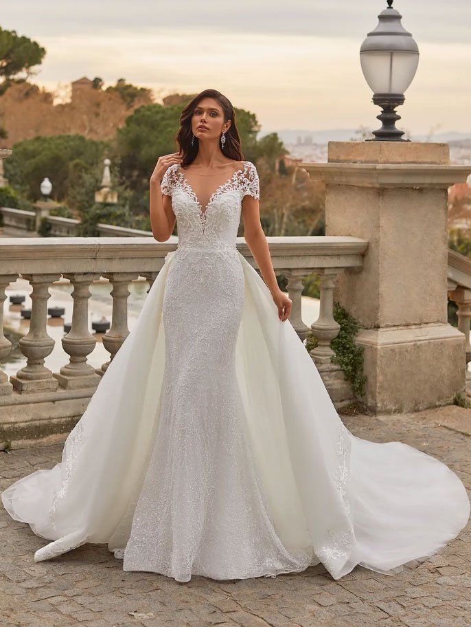 Una persona con indosso un abito da sposa di pizzo bianco, con sopragonna removibile e maniche corte, posa su un balcone di pietra al tramonto.