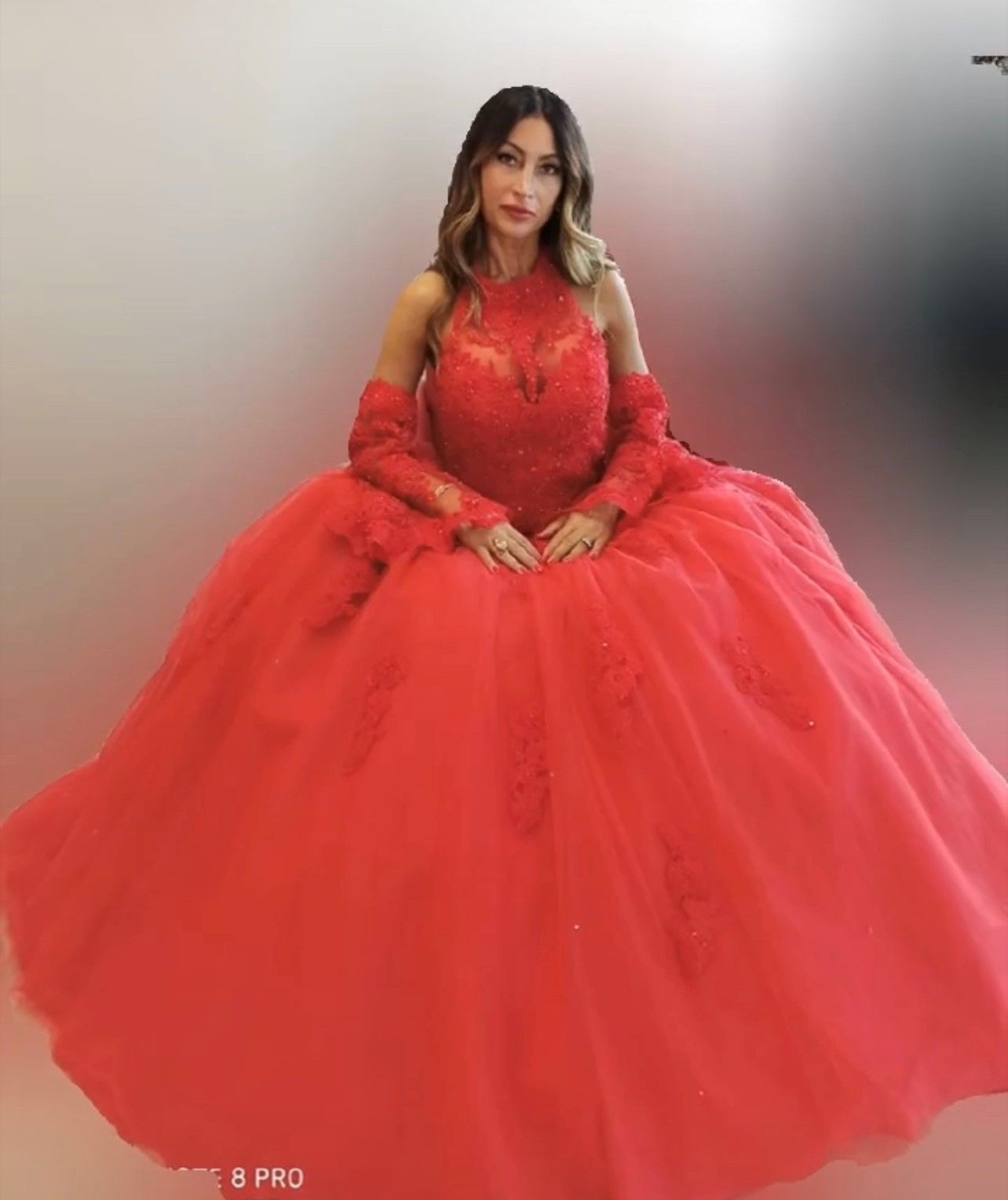 Una persona che indossa un abito da ballo rosso acceso, lungo fino ai piedi, con maniche lunghe e dettagli in pizzo, posa davanti a una parete spoglia.