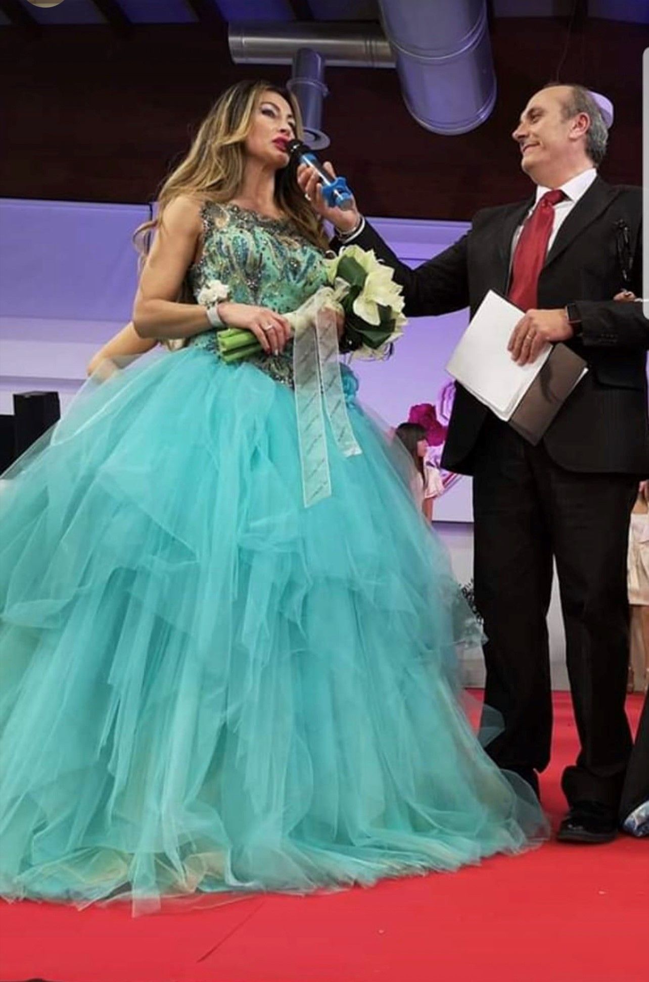 Una donna con un voluminoso abito da sera color verde acqua, che tiene in mano dei fiori, parla al microfono accanto a un uomo in giacca e cravatta che regge una cartella.