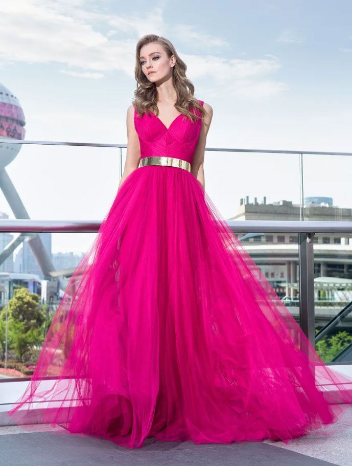 Una persona che indossa un abito lungo fino ai piedi color magenta acceso con una cintura dorata metallizzata, posa su una terrazza panoramica all'aperto.