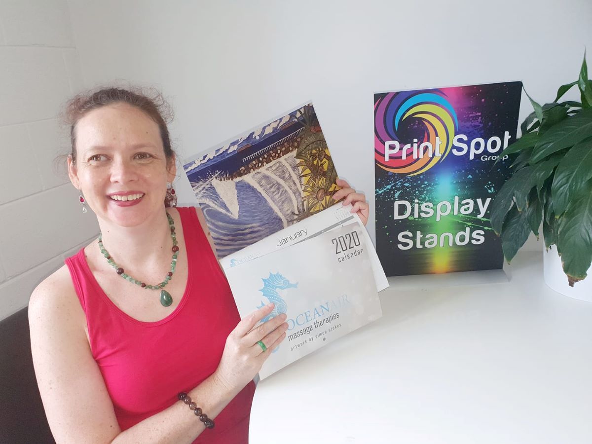 Woman Is Holding A Calendar In Front Of A Sign that Says Display Stands — Print Spot Group in Byron Bay, NSW
