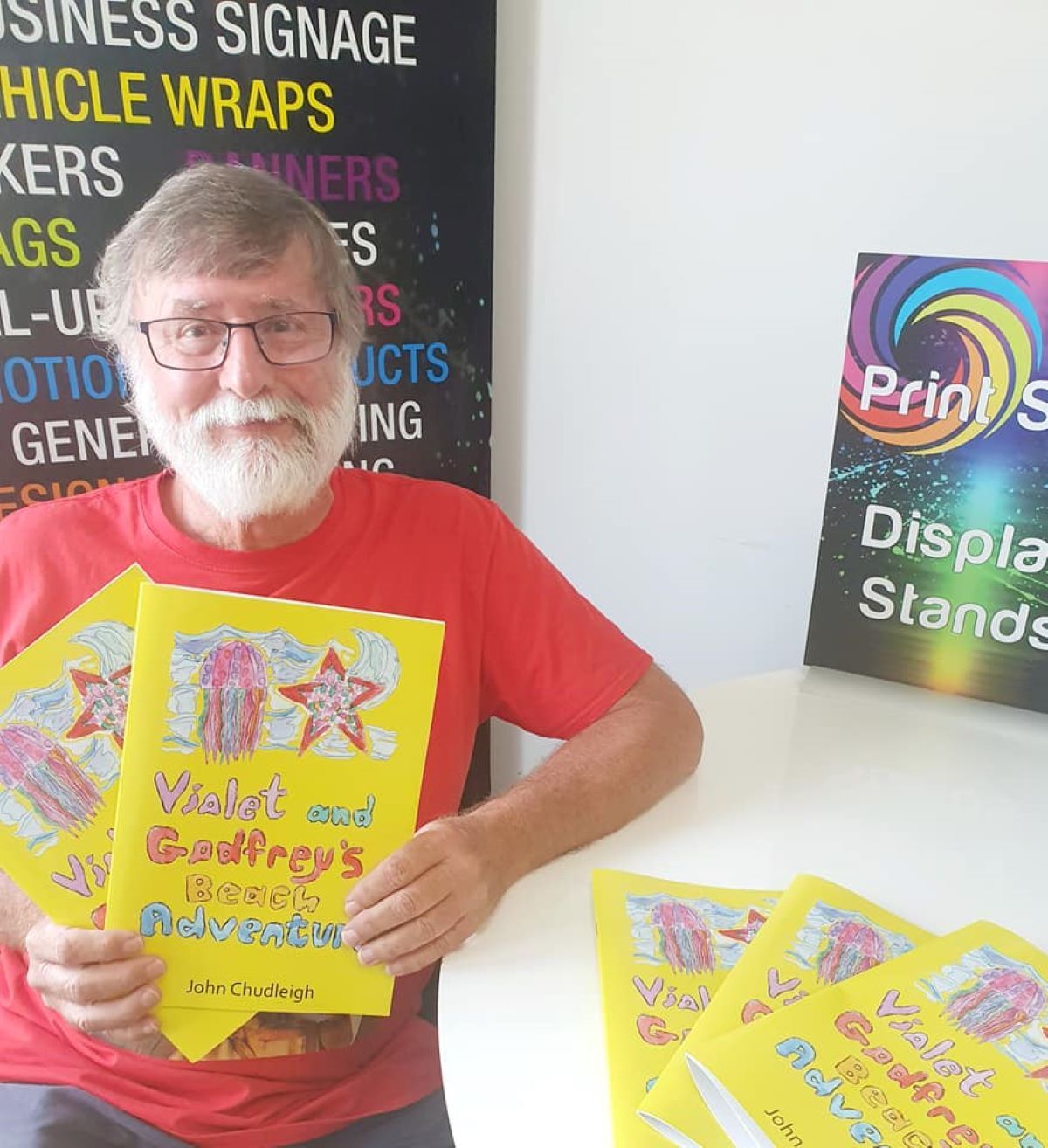 A Man in a Red Shirt is Holding a Book in Front of a Sign — Print Spot Group in Kingscliff, NSW