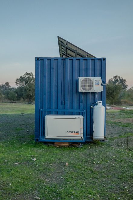 Blue shipping container with solar panel, air conditioner unit, and propane tank in a grassy field at dusk
