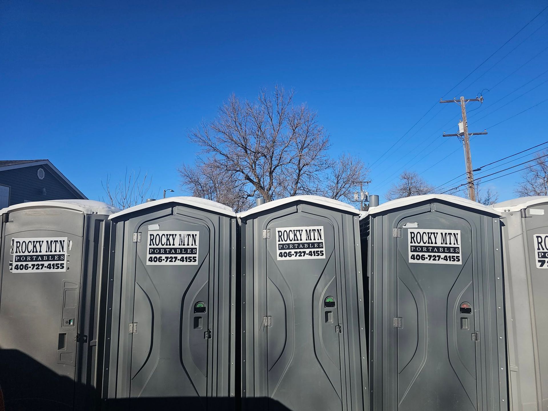Row of gray portable toilets under a clear blue sky beside a house and bare tree