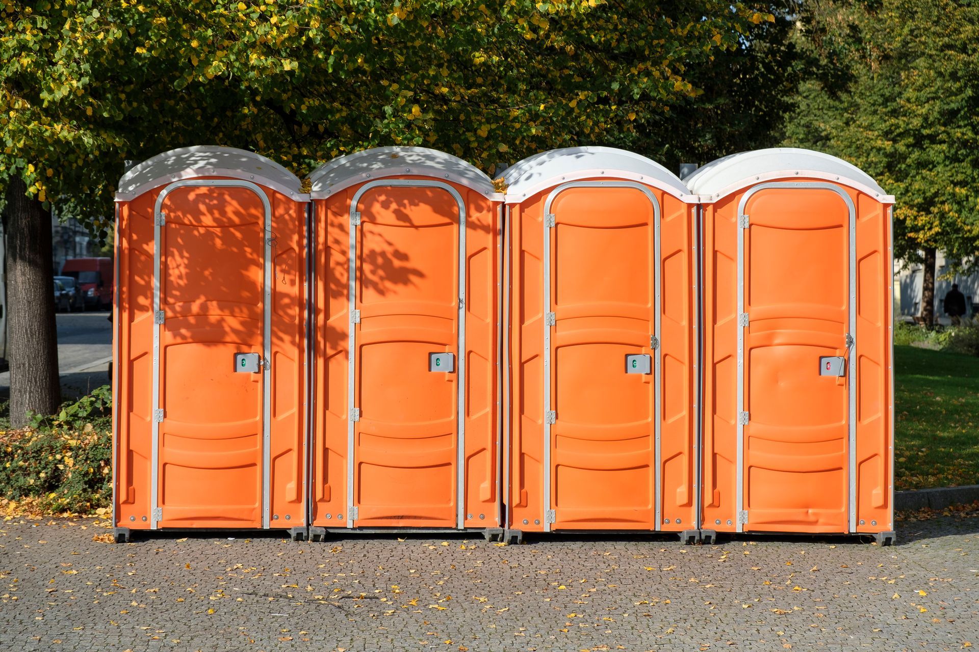 Four orange portable toilets lined up outdoors on a gravel lot under trees.