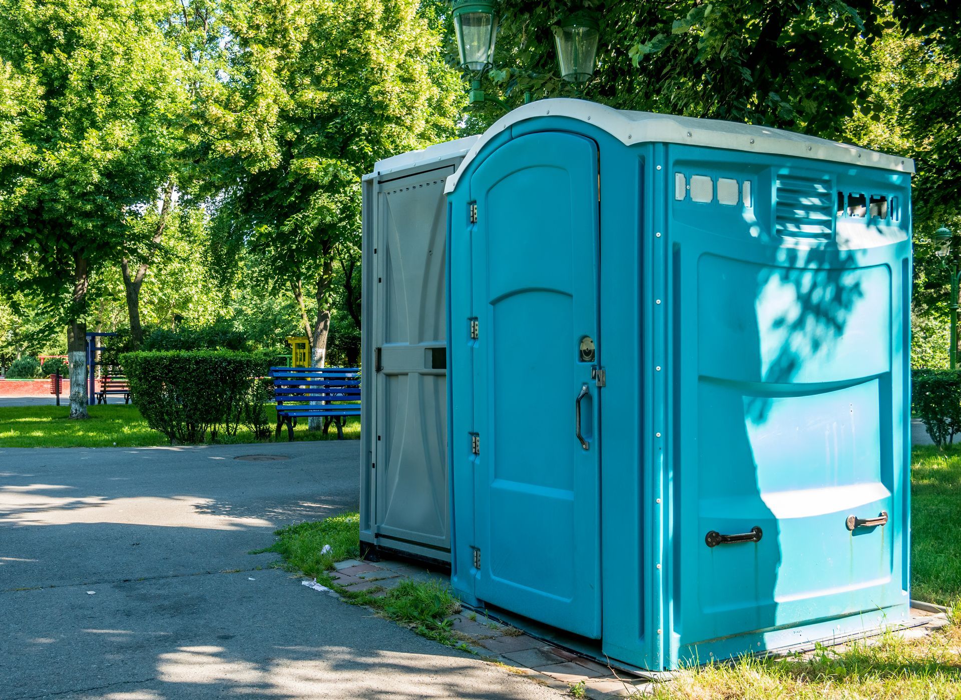 Blue portable toilet beside a gray portable shower in a sunny park