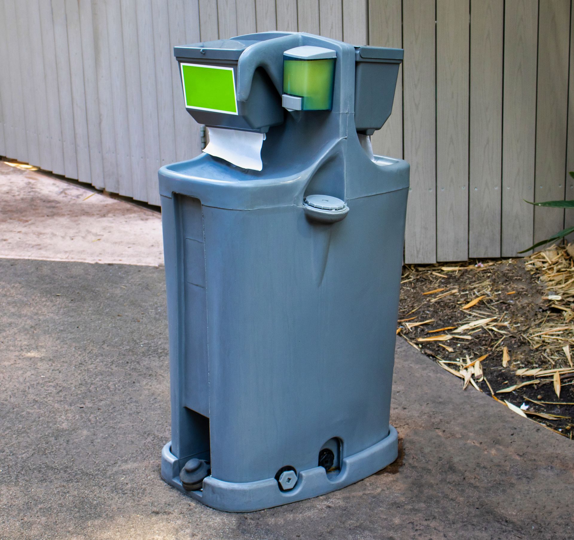 Blue-gray trash can with bright green panels on a sidewalk beside a wooden wall.