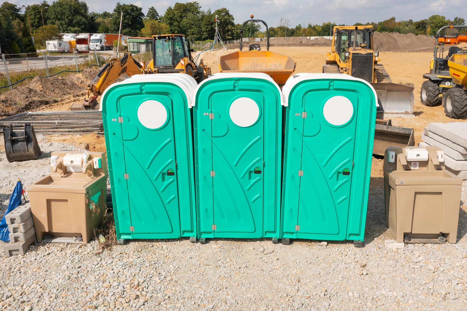 Three turquoise portable toilets at a construction site, with excavators and dirt in the background