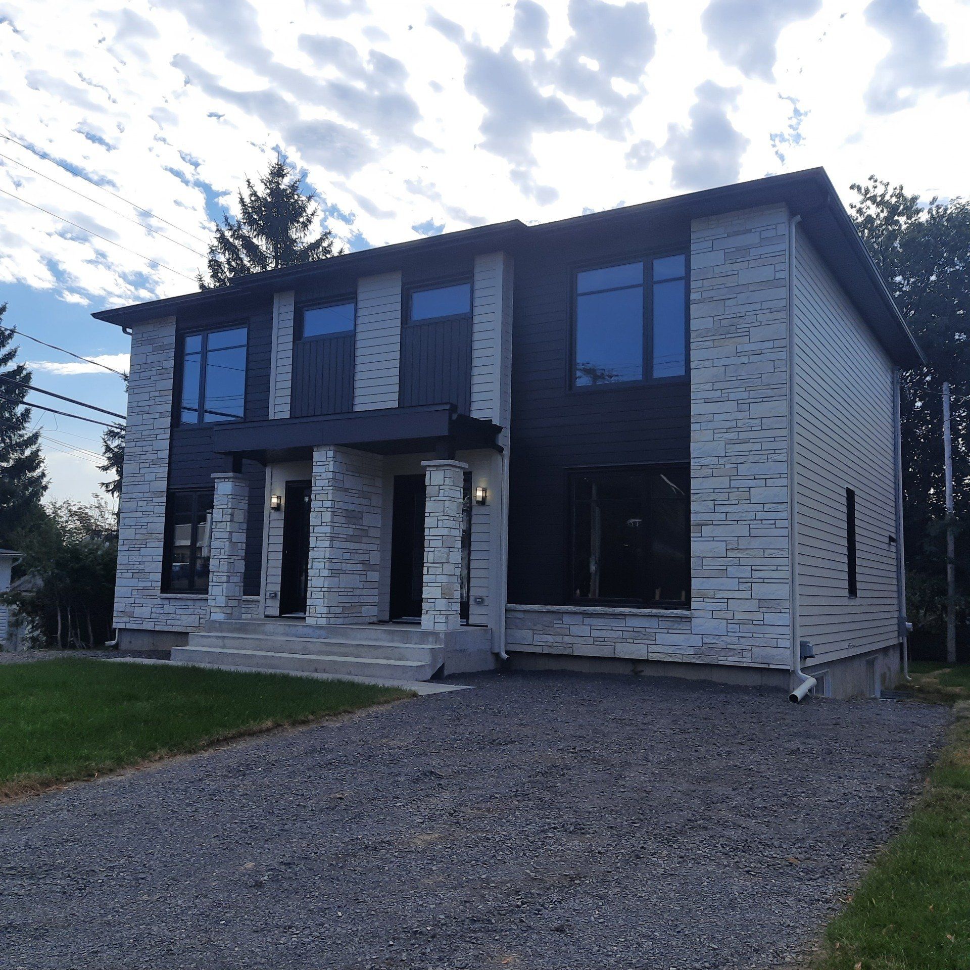 Duplex moderne avec revêtement gris et noir, allée en gravier, sous un ciel nuageux.