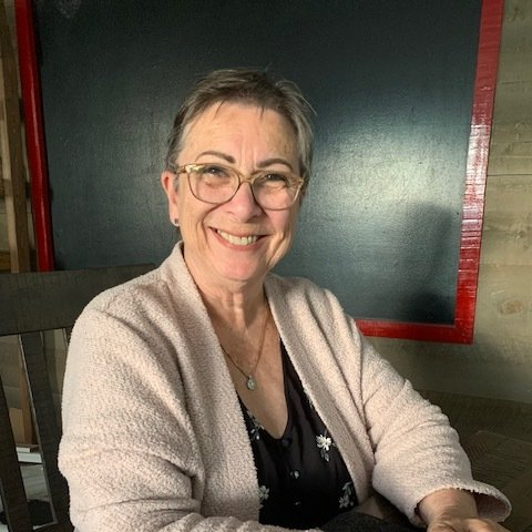 Femme souriante avec des lunettes, des cheveux courts et un pull rose clair, assise à l'intérieur.