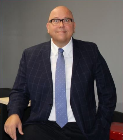 Homme chauve à lunettes, blazer bleu marine, cravate bleu clair, souriant, appuyé contre un mur gris.