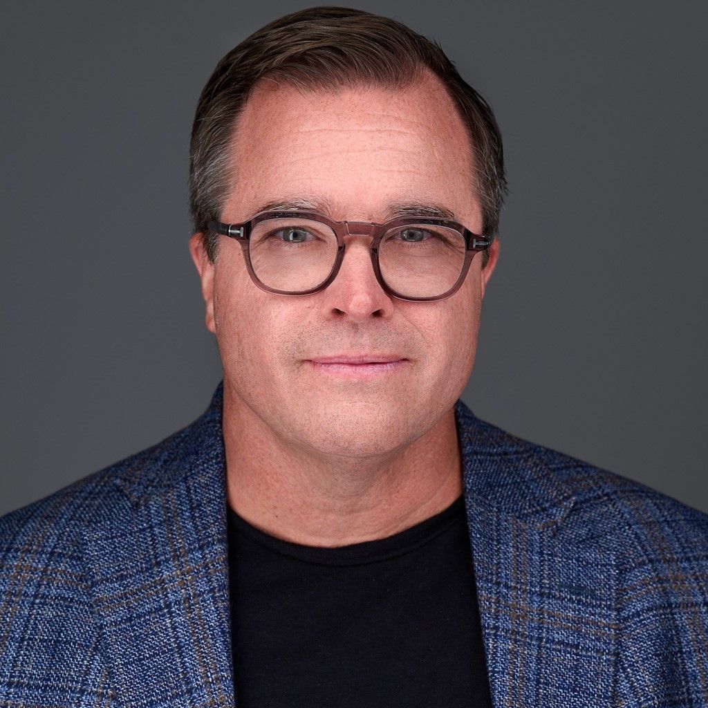 Homme avec des lunettes, portant une veste bleue et une chemise noire, expression neutre, fond gris.
