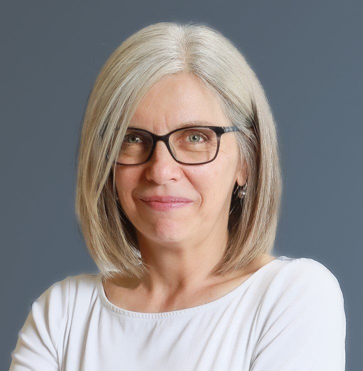 Une femme aux cheveux gris, portant des lunettes et un haut blanc sourit à la caméra, sur un fond gris.