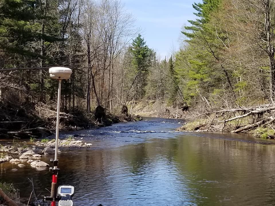 A survey GPS receiver on a tripod positioned at the edge of a forest stream on a sunny day.