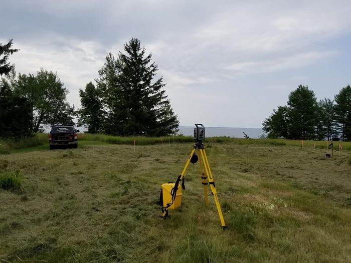 A yellow survey tripod stands in a freshly mowed field with a dark vehicle parked near trees in the background.
