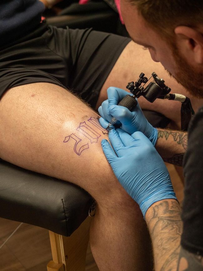 A tattoo artist wearing blue gloves uses a rotary tattoo machine to outline a gothic-style number on a person's thigh.