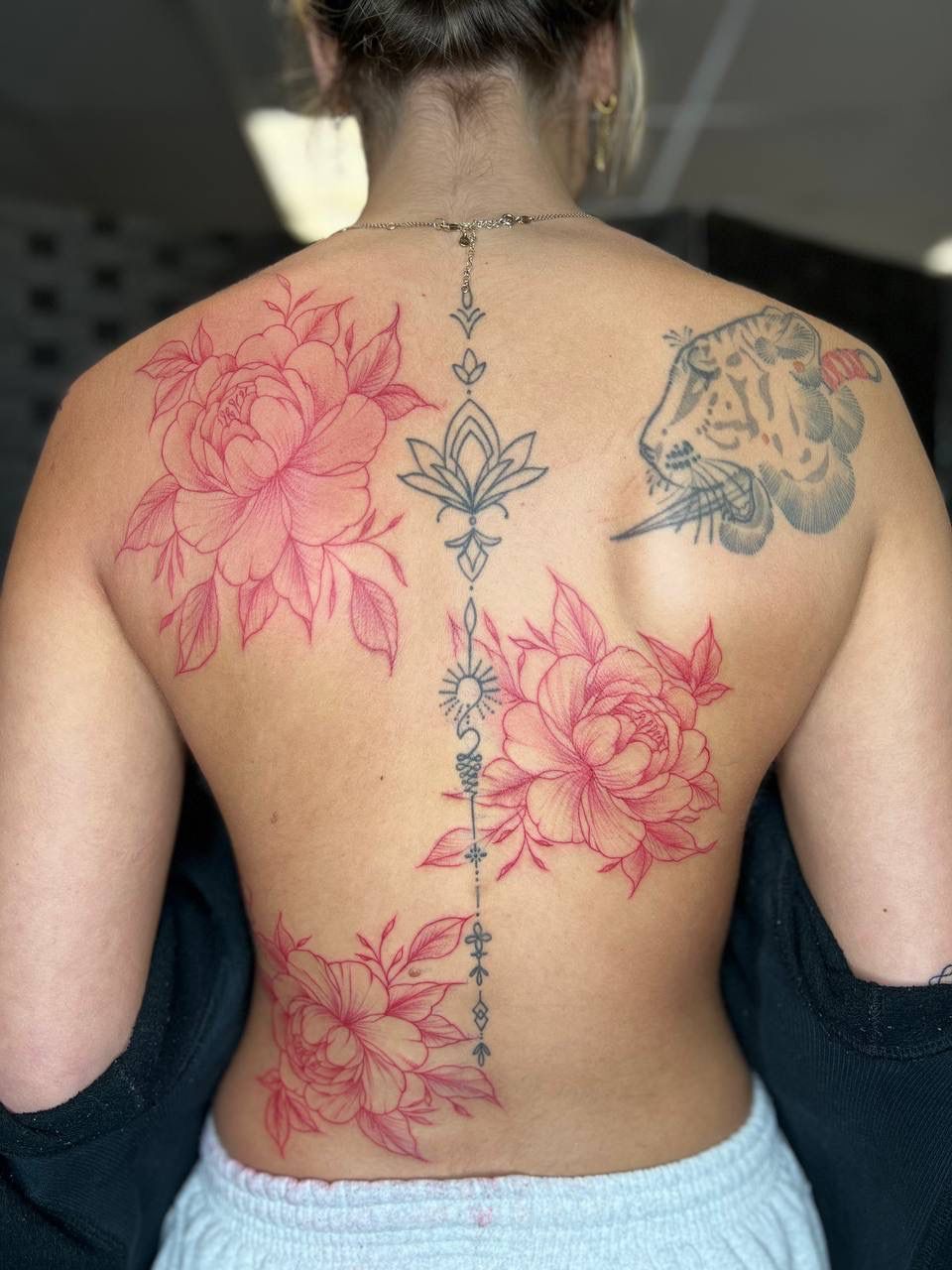 A person's back displays red floral tattoos surrounding a black linear spine piece, plus a tiger tattoo on the shoulder.