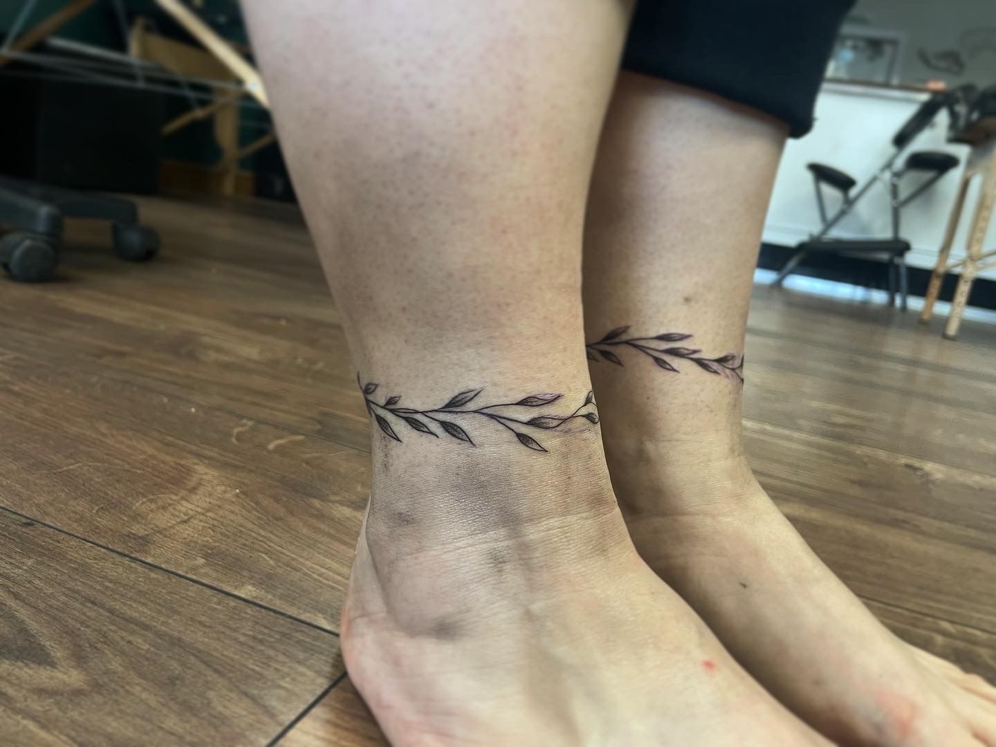 A pair of ankles each featuring a matching black ink, vine-style tattoo encircling the lower leg.