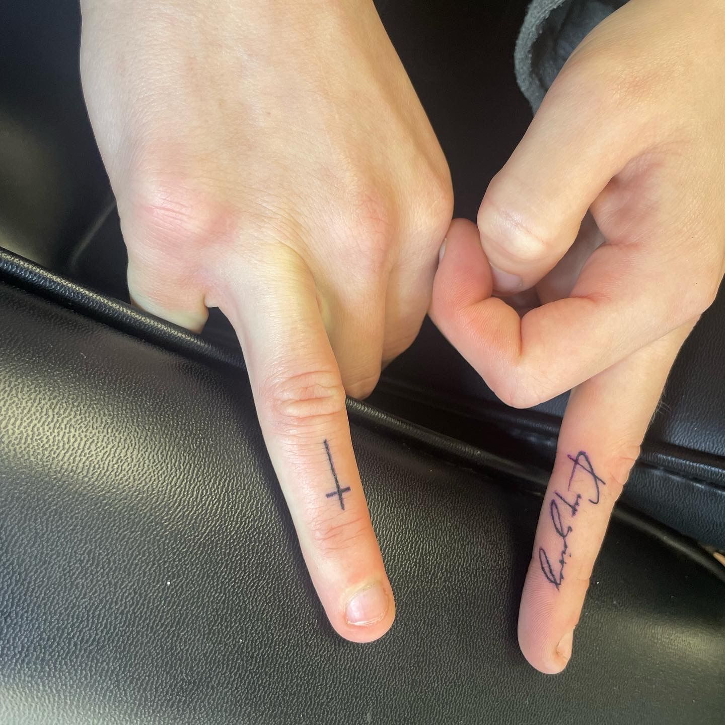 Two fingers with tattoos: one shows a thin, vertical cross, and the other features script lettering.